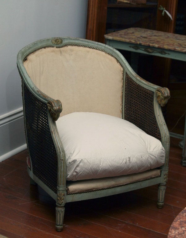 A painted Louis XVI style doubled caned chair with a loose cushion, each arm terminating in a ram's head.