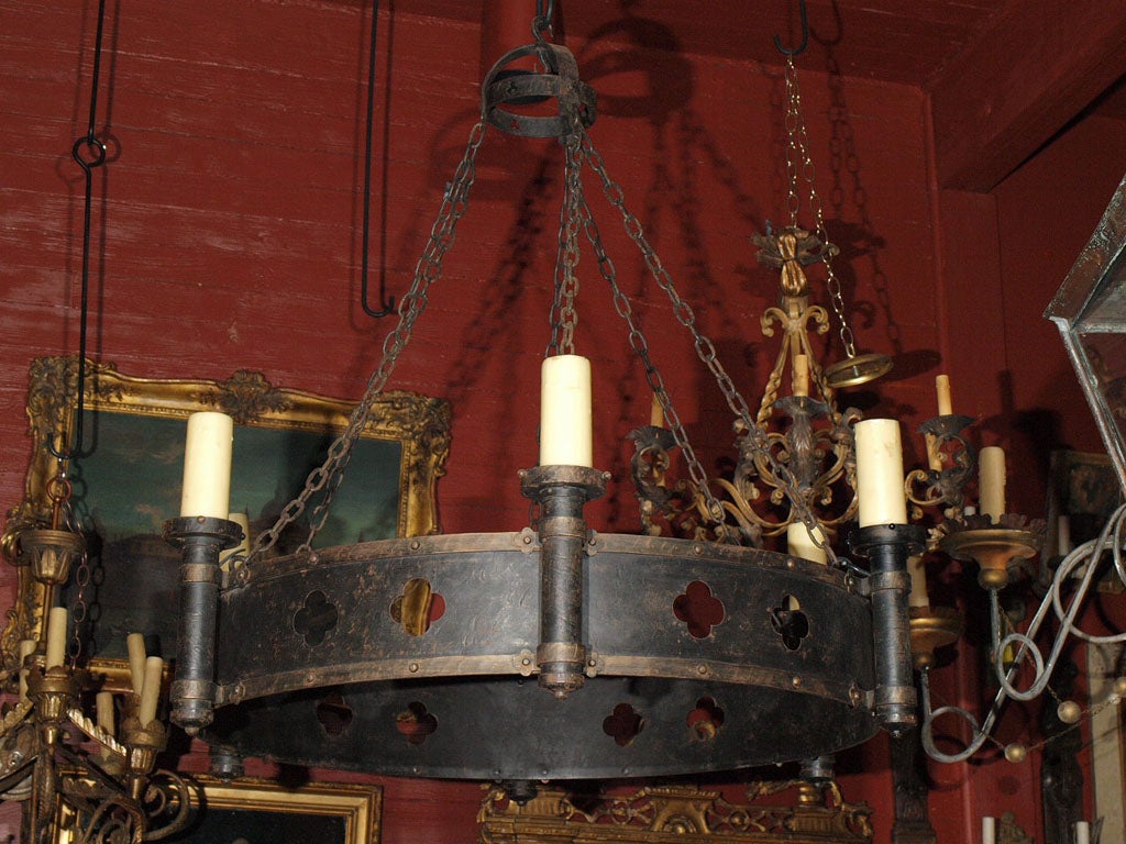A large black iron round chandelier. A pierced band with quatrefoil with dark gold banding. Three sets of heavy chain attach the ring to the canopy.
US wired