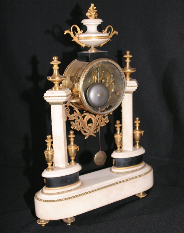 THE CYLINDRICAL CASE WITH ENAMEL DIAL AND ORNATE APRON, SURMOUNTED BY AN URN WITH FLORAL FINIAL, SUPPORTED BY SQUARE COLUMNS ON OVAL STANDS, THE WHOLE RESTING ON AN OBLONG BASE RAISED ON TOUPIE FEET, FITTED WITH GILT-BRONZE URNS AND DECORATIVE
