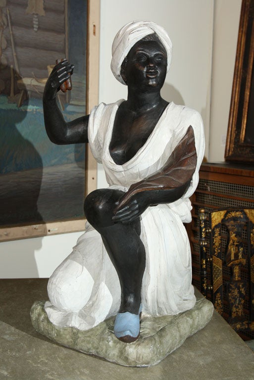 An English or American elegantly carved and polychrome wooden figure of a Negro slave woman clothed in a white sheath dress and matching turban holding a tobacco leaf and a bunch of cigars.