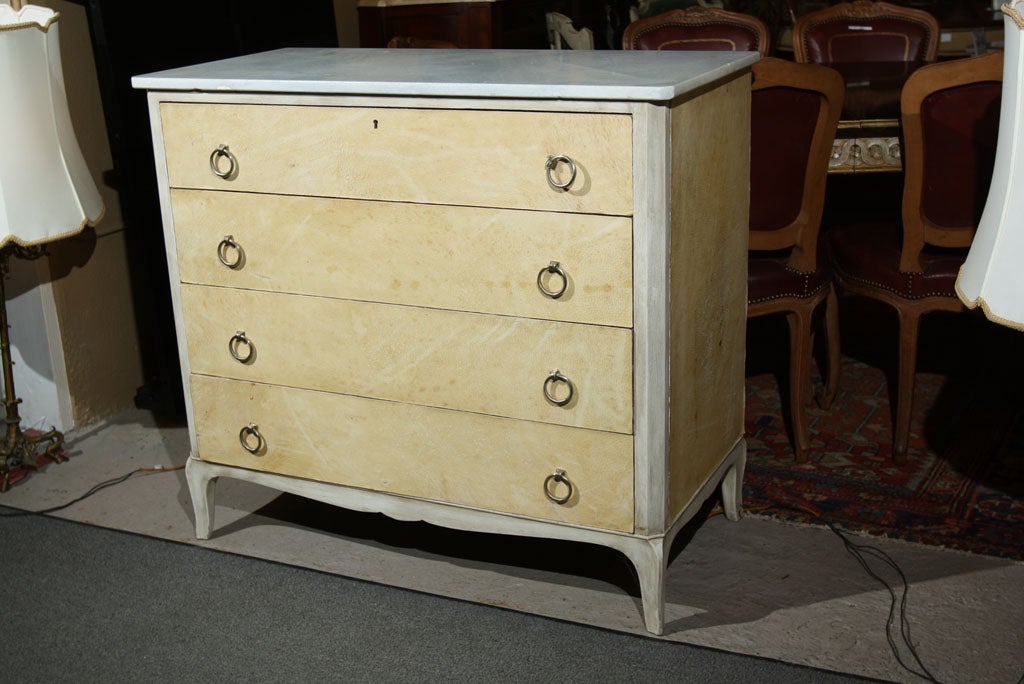 An intriguing French chest of drawers, the white marble atop a creme peinte conforming case of four drawers, veneered with parchment, the top drawer opens to a writing surface with cubby holes, standing on cabriole legs. From our Winter White