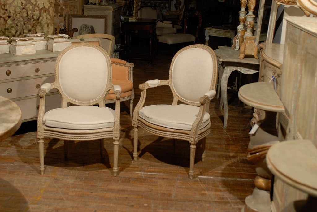 A pair of French Louis XVI style oval back painted wood upholstered bergeres chairs from the 1920s-1930s. This pair of French bergeres chairs have an upholstered oval back topped with a rose and ribbon carving. The scrolled arms have partial