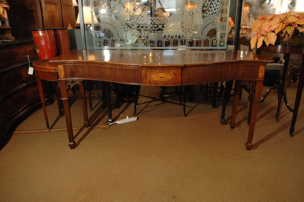 A George III serpentine front sideboard table of mahogany with fluted carved apron, inlay, and trapezoidal legs.