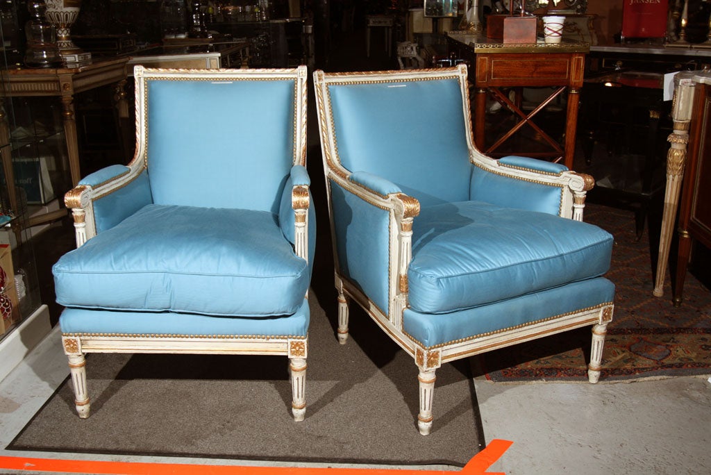 A white painted and parcel-gilt French bergere chair with blue silk upholstery, in the style of Louis XVI, stamped Jansen. This finely upholstered chair in a white and gilt gold decorated frame with intricate carvings would make a fine addition to