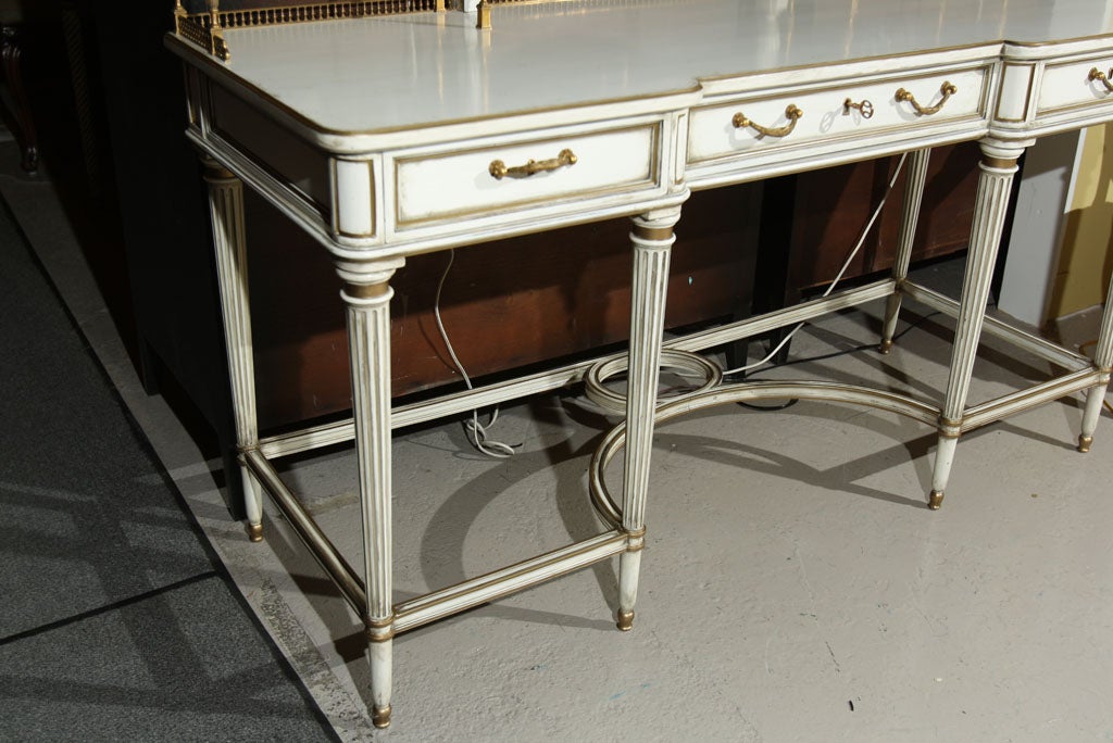 A charming vanity, circa 1930s, thoroughly white painted and parcel-gilded, the oval mirror flanked by bronze galleried trays, supported by a frieze of three drawers, standing on fluted tapering legs ending in capped feet. STAMPED JANSEN.