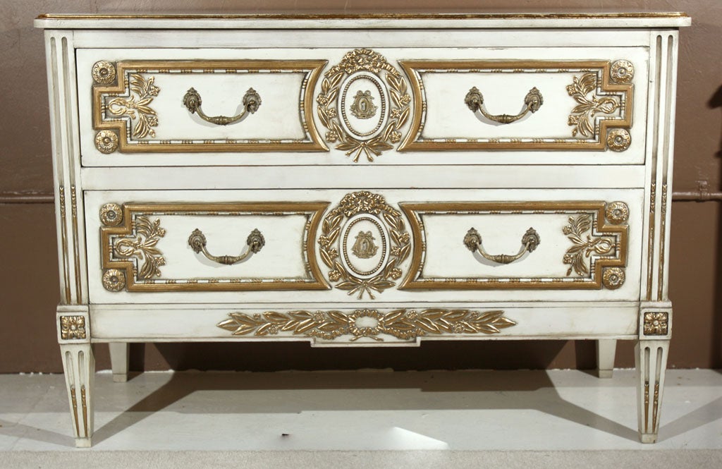 A very unique two-drawer commode, circa 1940s, thoroughly creme peinte and parcel gilded, the rectangular beveled top over a conforming case of two drawers, elegantly carved medallion, wreath, and floral patera decorated with ormolu and gilded