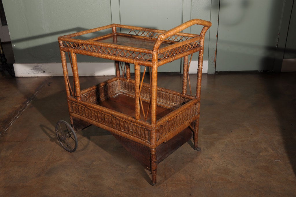 An Edwardian-era drinks cart or trolley from England featuring two wooden tiers with woven wicker galleries, set upon a wood and woven wicker framework and resting on spoked wheels and casters.<br />
<br />
Used for tea, desserts, and beverages,