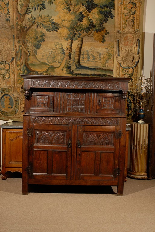 English oak court cupboard with carved frieze and finials above  enclosed cabinets with carved doors and iron hardware. The central panel with carved ca.date. Below are two large  cabinets with shelf. All resting on raised legs. <br />
<br />
For