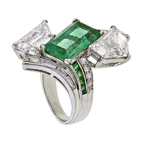 This dynamic, large scale, adaptation of the classic platinum by-pass ring is designed around a vivacious, bright green, elongated emerald-cut emerald weighing 4.00 carats. The emerald is asymmetrically framed by two large fancy-cut diamonds: an