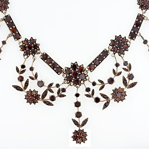 This early 20th century necklace is made in the Bohemian garnet style of the late 1800's. It is studded with glowing orangy red garnets and draped in festoons of floral garlands. The garnets are set in rose gilded base metal.  17 3/4 inches in