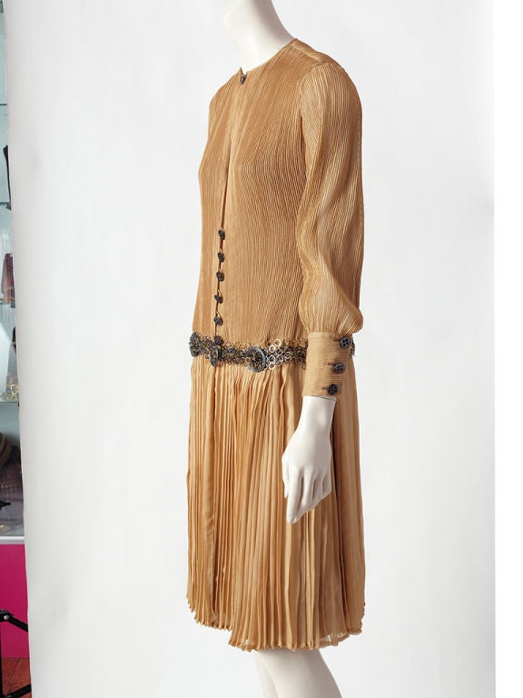 James Galanos beige, silk chiffon, dropped waist day dress. Bodice  and long sleeves have vertical tiny, fine, pin tucking detail. Skirt is pleated. Round neckline closes with a pewter toned button and is open until the center of the chest, where