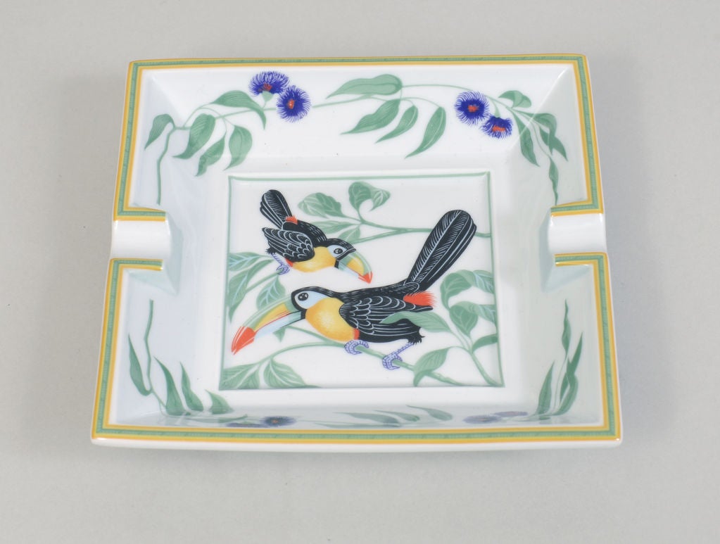 A decorative porcelain ashtray from Hermes, Paris is still in the original box.  The Toucan pattern has two colorful birds in the center surrounded by flowering vines.  The base is covered with green suede and marked Hermes, Paris in gold.  The