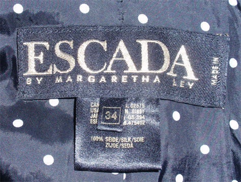 This is a black sequin trench coat style coat by Escada, from the 1990's. The coat is knee length with two front flap pockets, a large collar, and shoulder pads. It has an adjustable belt with a black, white polka dot silk lining, 20.5