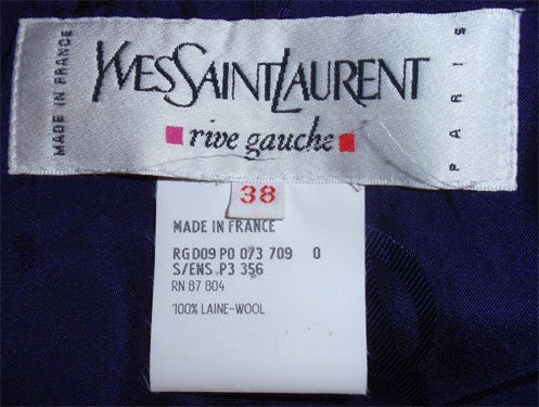Si tratta di un completo di giacca, camicetta e gonna di Yves St Laurent, risalente agli anni '90. La giacca e la gonna sono in lana leggera con una stampa a righe viola. La camicetta è in seta con maniche lunghe e bottoni sulla