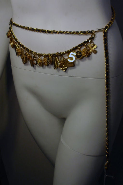 This is a gold charm belt by Chanel, from the 1990's. It has a black leather in the gold chain. The belt is adjustable in size, and the charms are all icons of Chanel.<br />
This gold and black charm belt by Chanel is available to be viewed