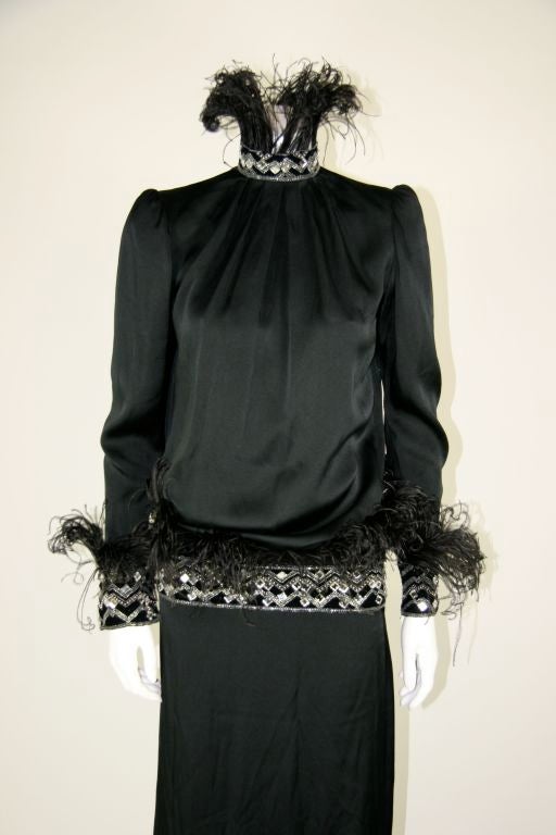 Givenchy black silk jersey gown with full length sleeves. Velvet trim on neck, wrists and hip with square chisel cut rhinestones, silver beading and ostrich feathers sewn upward. Open back from neck to the hip. Bodice blousons over the hip trim and