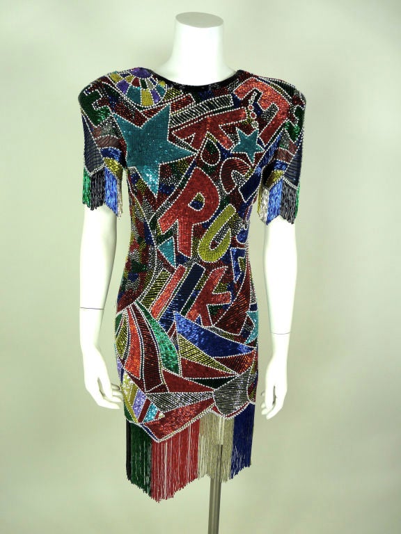 Fabrice multi colored beaded cocktail graffiti dress with fringes. Dress has low back with short sleeves. Fringes on the sleeves and on the hem. Hem is serrated. Shoulder pads in the dress. Pattern is letters, stars and graffiti fan fare design. <br
