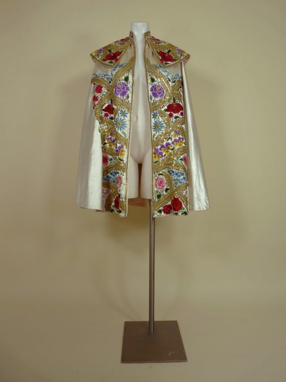 Circa 1940s (?) white satin hand embroidered cape from a Suit of Lights matatdor costume. Cape is embellished with vibrantly colored floral motif embroidery and gold bullion foliate with sequined embroidery. Lined in pale pink satin. Made in Madrid,
