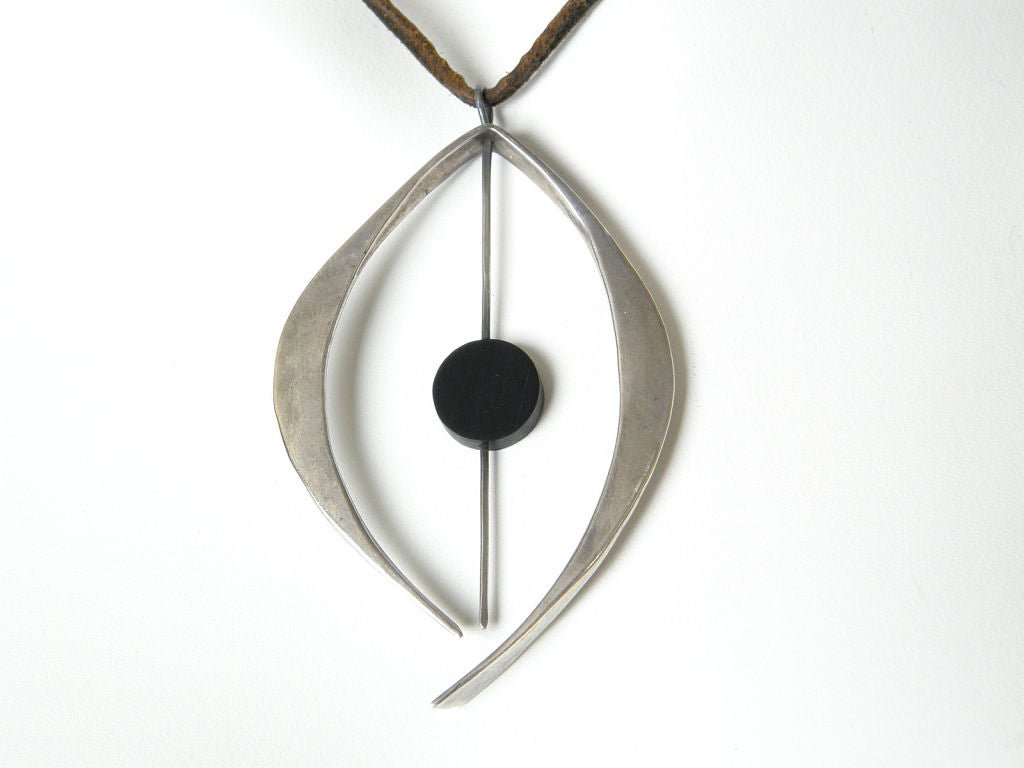 This necklace is a wonderful example of Betty Cooke's earlier work. It has a two part kinetic pendant with a flat ebony disk skewered on a sterling rod and bracketed by the curving, hammered piece. It hangs from its original leather cord which has a