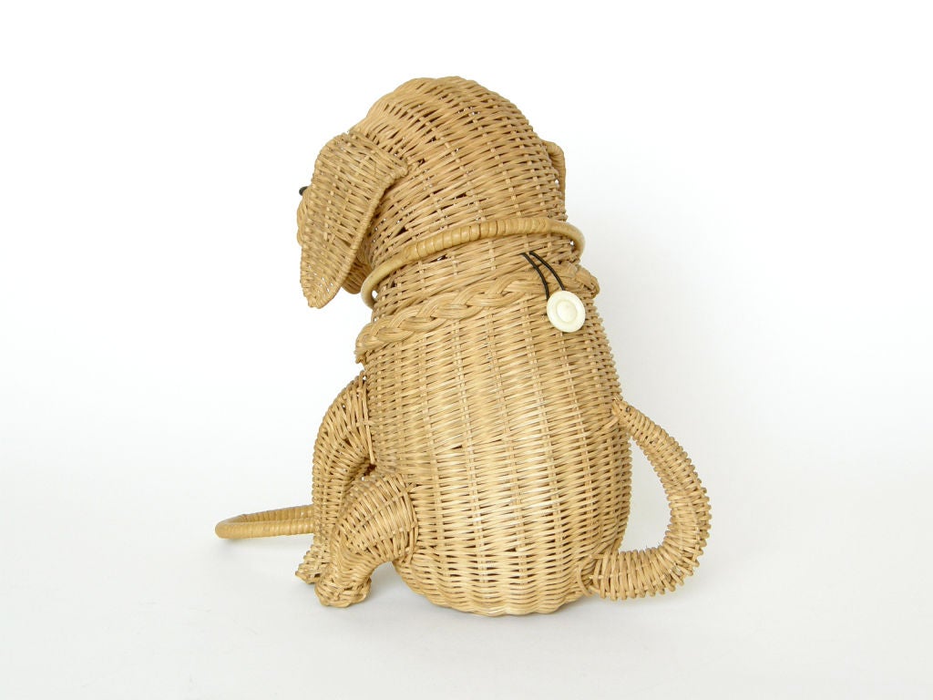 This sweet wicker handbag is in the shape of a seated dog. Her collar and leash form the handle. This bag has a charming quality with a pose and face that are almost realistic in a cartoonlike way. It opens at the neck with an elastic loop and