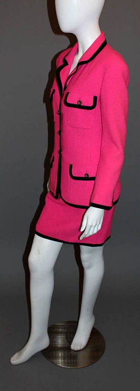 This spectacular skirt suit hails from CHANEL Boutique (Collection 25). Composed of bright pink boucle, the jacket features a rounded, notched collar, gold and black button down front, tapered waist and four pockets in front. Full-length sleeves