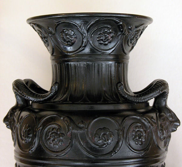 A striking pair of English neoclassical style Wedgwood jasper-ware urns with rams head handles; each with everted lip above a bulbous fluted body flanked by rams head handles with raised foliate scrolls at the neck; raised on a tapering base;