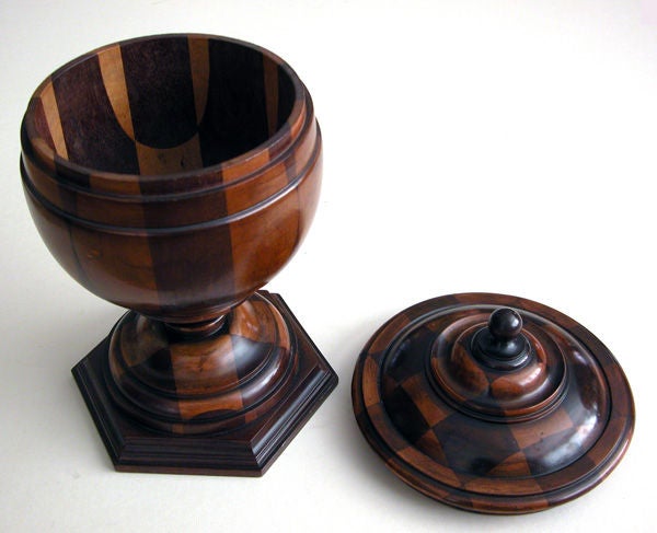 A warmly-patinated English turned-treenware covered urn; of contrasting woods, the domed lid with finial knob above a portly body above a stepped foot; all over a hexagonal plinth