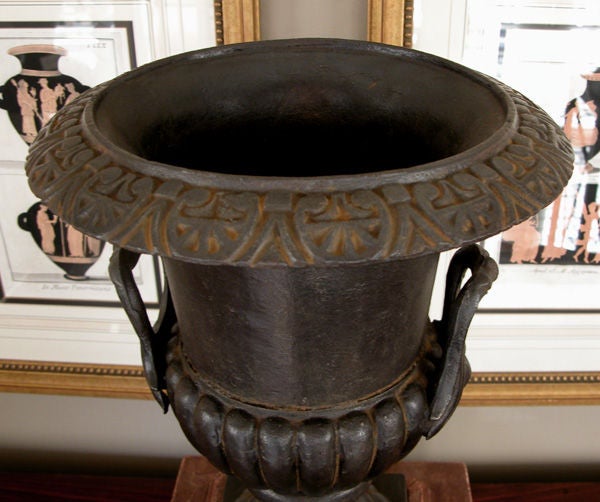 A handsome French baroque style double-handled iron campagna urn; the everted lip with leaf-and-dart motif above a compressed lobed body; raised on a flared base; the whole on a later faux marble painted wooden base
