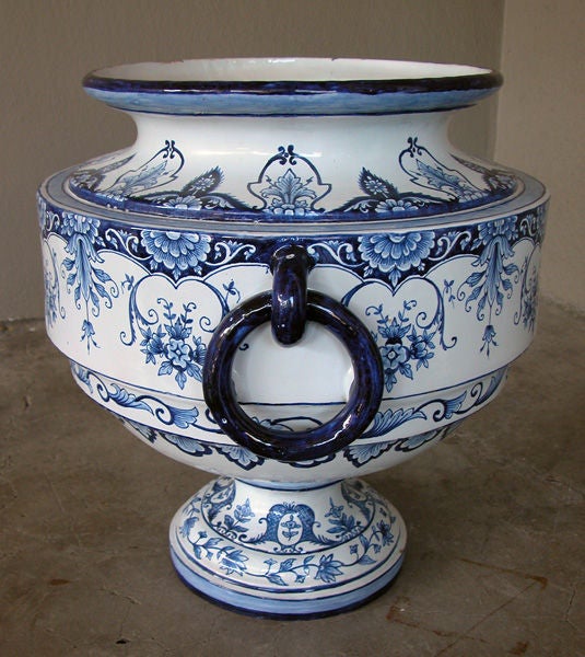 A massive French blue and white tin-glazed faience double-handled urn; with short neck above a compressed square body flanked by applied ring handles; raised on a flared circular base; painted overall with lively floral and foliate decoration