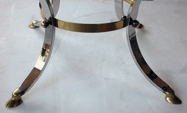 A stylish American 1960's chrome and brass circular cocktail table with glass top; the thick circular glass top resting on a circular ring raised on dramatically splayed supports ending in brass hoof feet