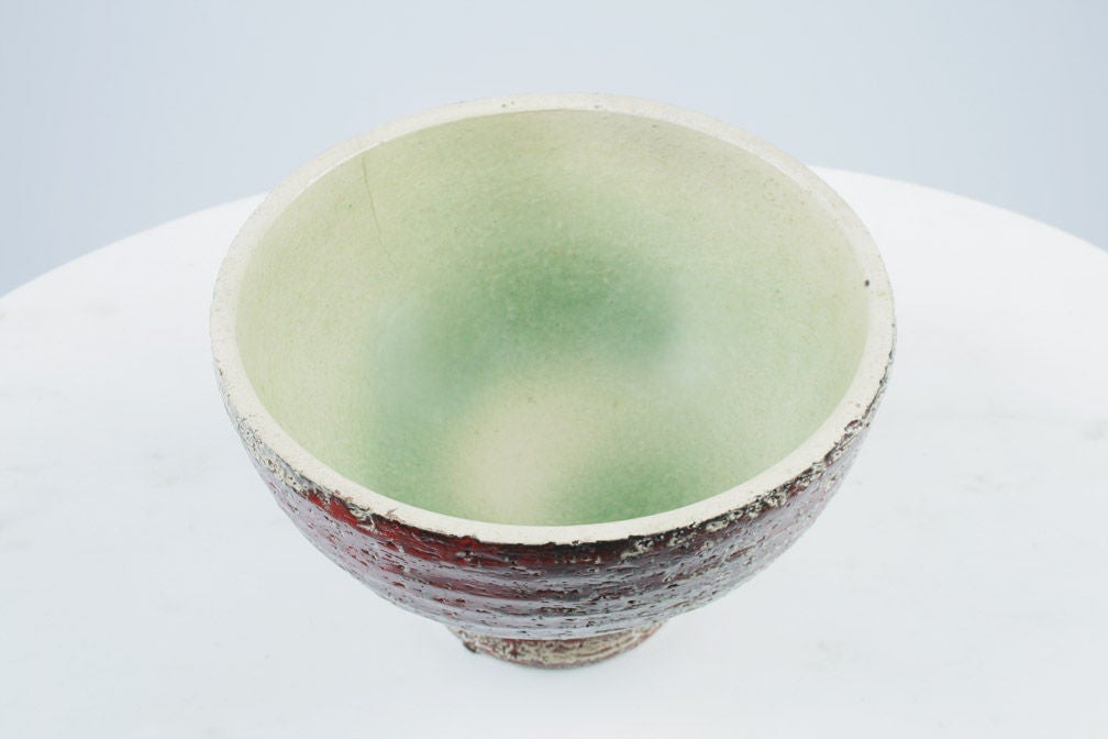 A vibrant compote consisting of a stout bowl with a textured, ribbed exterior in a red and cream glaze, a smooth interior with a celadon colored swirl glaze motif, all supported by a short flared foot.  Italian design imported by the New York based
