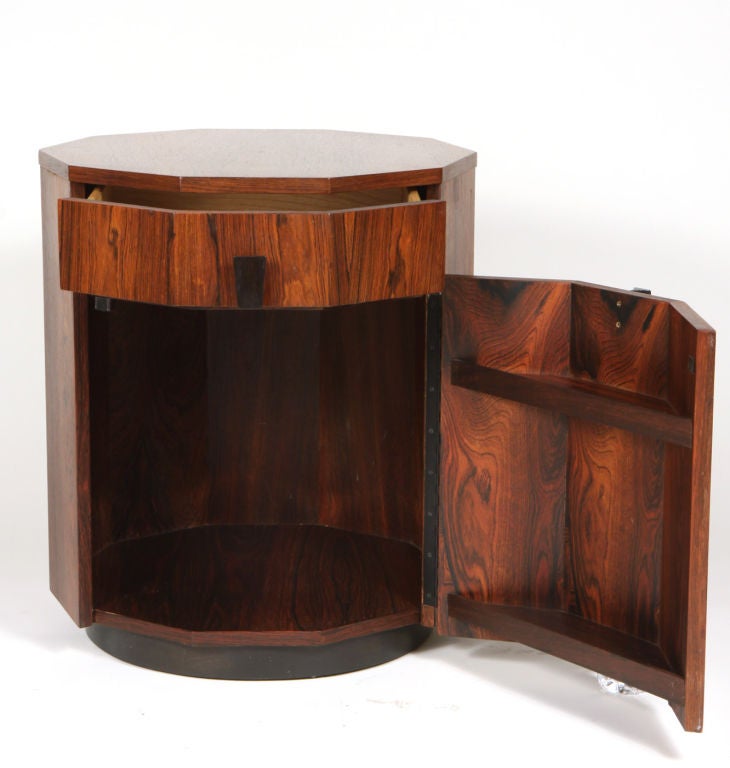 A ten-sided rosewood dry bar cabinet or lamp table with ebonized trapezoidal wood pulls. Upper drawer over door compartment and round wood plinth. Door incorporates two galleries for storing bottles. Signed with maker's label and burned in mark.