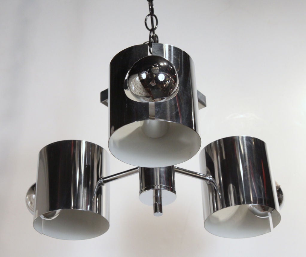 A mod polished chrome ceiling light with a square canopy that holds a short shaft with three arms, each ends in a cylindrical shade that houses an exposed clear silver bowl light bulb. American, circa 1970.