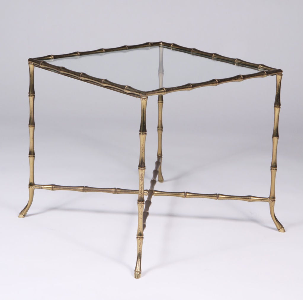 A chic 'Syrie' occasional table with an inset glass top and a square brass frame in a faux bamboo motif with an X-stretcher and petite flared feet. Manner of Bagues. By John Gerald, Inc.. U.S.A., circa 1950.