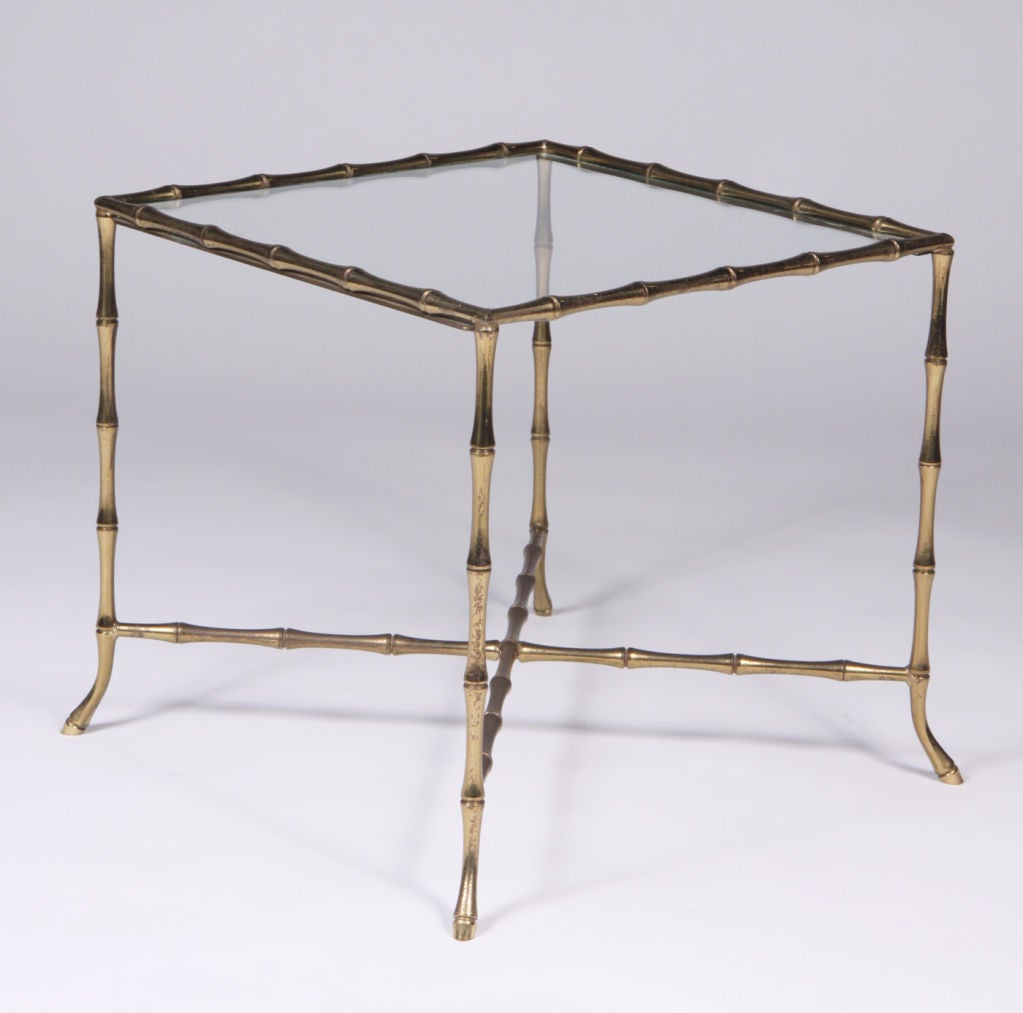 A chic 'Syrie' occasional table with an inset glass top and a square brass frame in a faux bamboo motif with an X-stretcher and petite flared feet. Manner of Bagues. By John Gerald, Inc.. U.S.A., circa 1950.