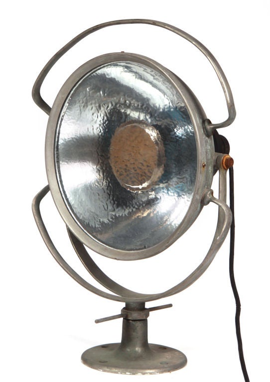 A vintage industrial search light with rounded handles attached to the top and bottom of the light, allowing up and down movement, the whole rests on a pedestal base that allows side to side movement. Metal stamp to the rear of the light.  By Wilmot
