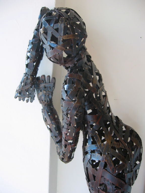 Contemporary sculpture fashioned in scorched metal and woven and soldered to create a human, female form. This is a three dimensional work of art which can be hung as if she were scaling a wall and it can also be placed in a position of repose. It