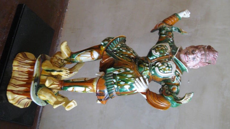 GLAZED MULTICOLOR TERRACOTTA MYTHICAL SCULPTURE. THE IMAGE OF A NOBLE WARRIOR REPRESENTING A GOD,DRESSED IN A COAT, PANTS AND BOOTS WITH INTRICATE DETAIL. THE FIGURE IS STANDING ON A SMALL MAN WHO REPRESENTS EVIL.