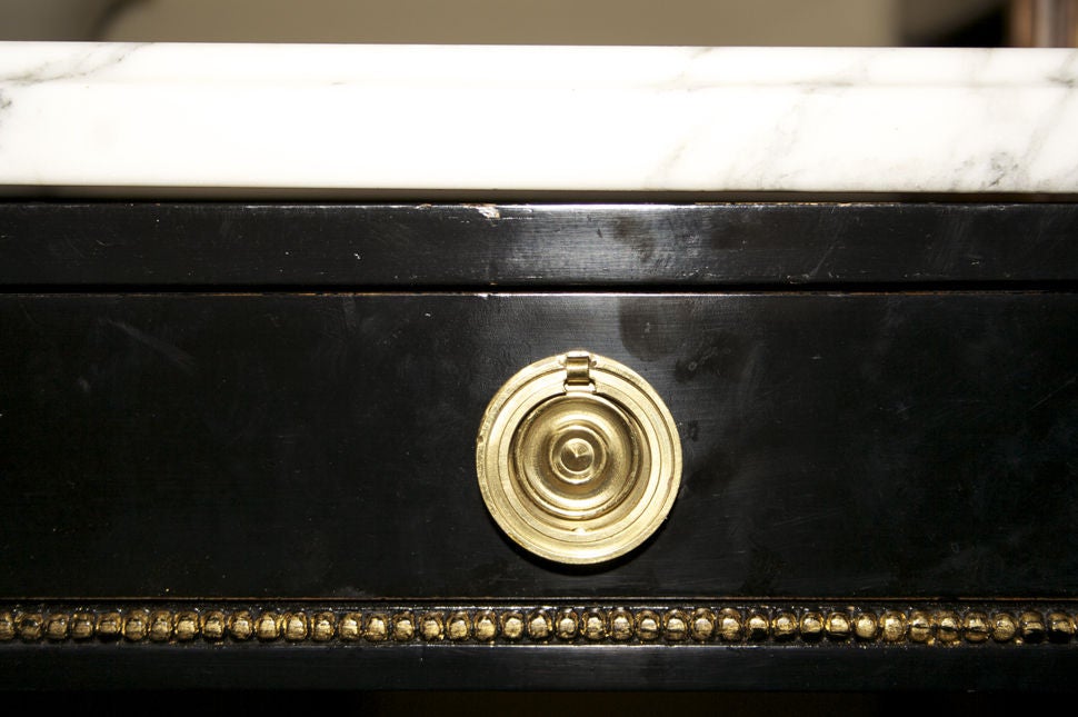 A French Louis XVI console table with four round fluted tapered legs joined by a stretcher with center rosette. The black painted frame having three drawers in its apron with brass pulls and marble top with ogee edge.