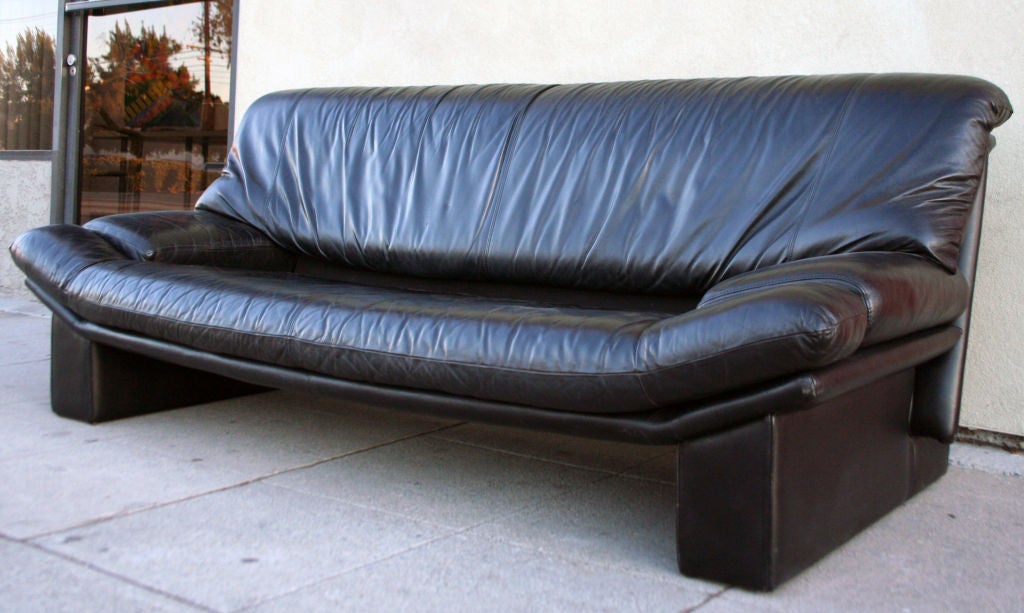 A Ligne Roset sofa with deep sinking seamless black leather backrest and seat and slightly tough armrest rolled over the seat cushion and into a double pedestal base which is also clad in black leather.