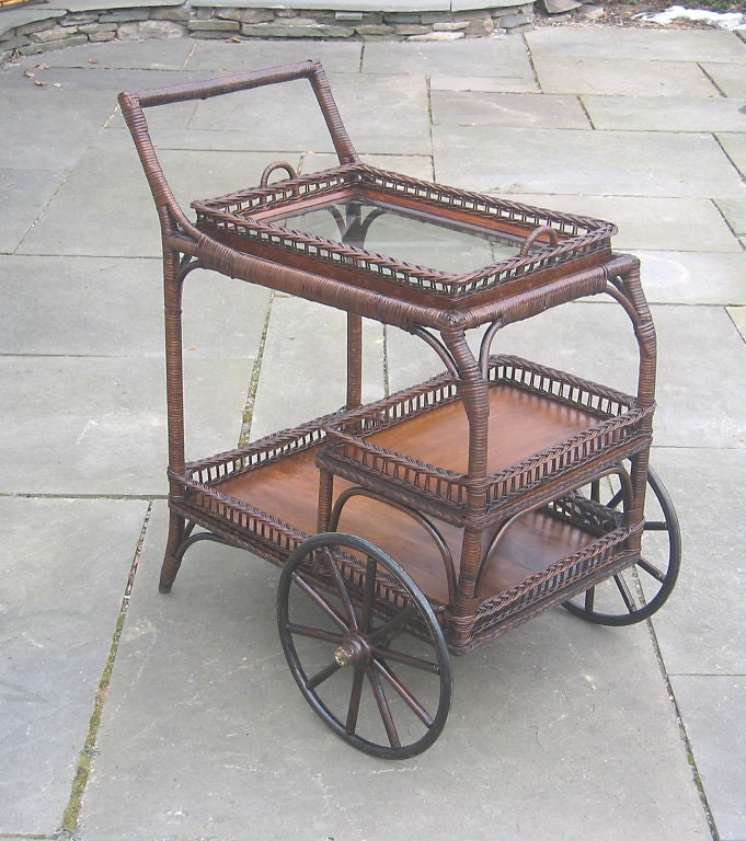American antique wicker tea cart in natural stained finish.  Full bottom shelf and center half shelf with wood surface.  Removable wicker framed glass serving tray with handles.  One & a half inch woven galleries having evenly space double reeds