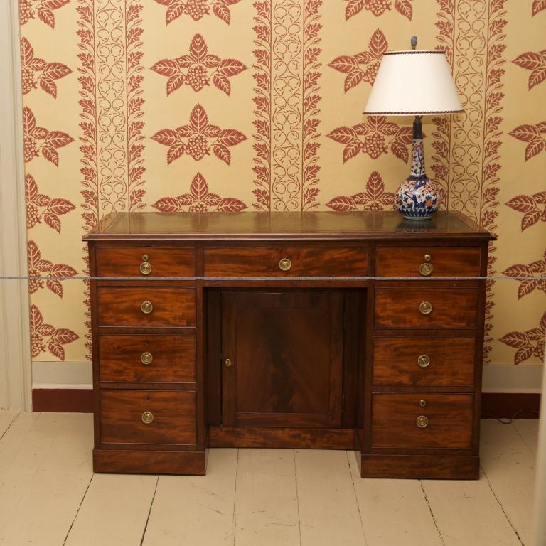 The narrow rectangular top with gold tooled green leather panel above 9 drawers, the knee hole front with cock beading and brass ring drop handles centering a recessed door, raised on a plinth.