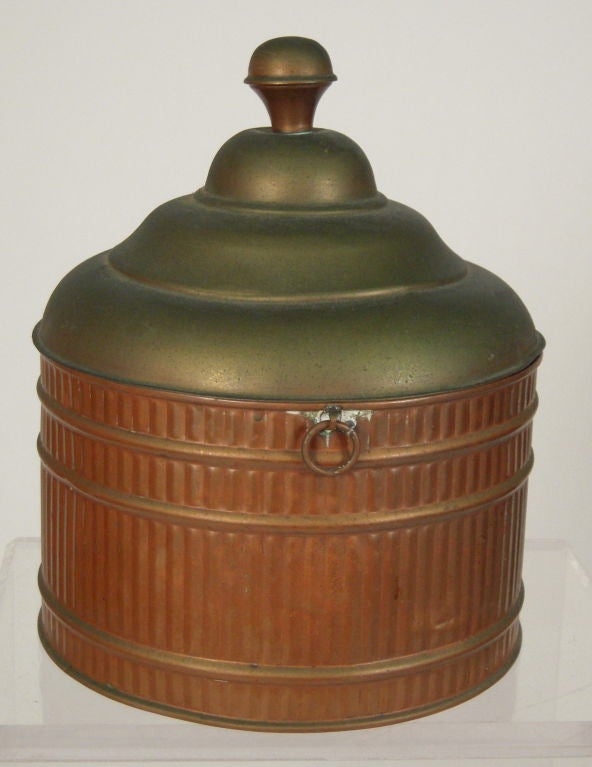 A round ribbed copper vessel with ring handles and a stepped, brass colored copper domed top with circular knob finial. Functional and decorative.