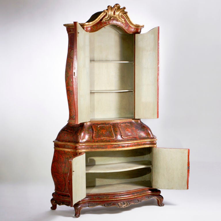 Venetian gesso polychrome and parcel-gilt display cabinet or bookcase decorated in red-orange colors. The arched feather pediment over a pair of molded doors open to a shelved interior. The center with small serpentine shaped paneled drawers. The