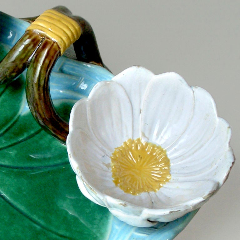 A large water lily leaf shaped Majolica strawberry server, strewn with strawberry blossoms and edged in blue, is modelled with two water lily flower cups for crème and sugar, handle with raffia tied centre.