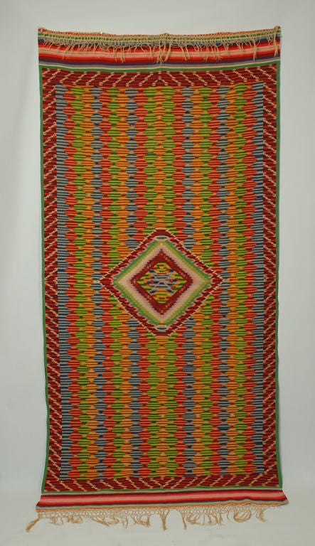 A graphic antique Mexican saltillo serape with a brilliant eye dazzler surrounded by a fully saturated palette of red, yellow, green, blue, pale blue and black diamond motifs and multiple striped borders. Incredibly tight weave with original hand