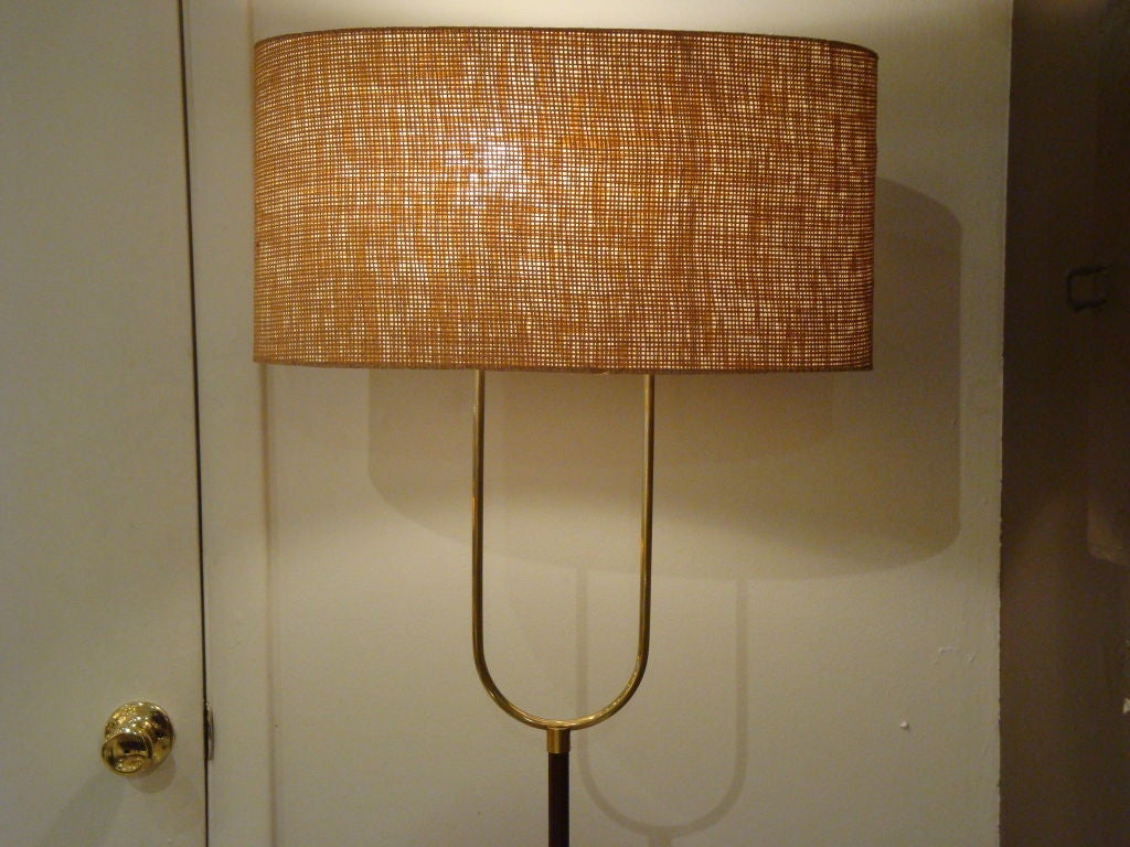 A modernist two light floor lamp featuring a wooden pole body, round weighted base in polished brass, a U shaped brass light support and a oval lamp shade in a heavy natural texture. France circa 1960.
