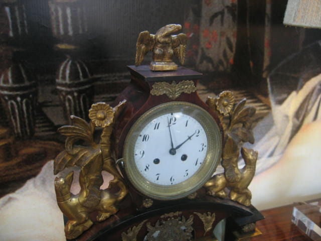 Beautifully designed mantle clock with ornate bronze embellishments. Carved and gilded griffins flank the clock face. A gilt eagle is the finial. The beautiful wood case is embellished with painted geometric designs and marble pilasters with gilded