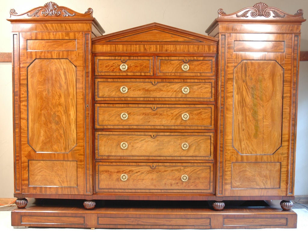 George IV part-ebonized mahogany wardrobe.  The center section with a triangular pediment above two short and four long graduated drawers, flanked by a pair of oversized cupboards surmounted by a scrolled cresting carved with palmetted and leaves