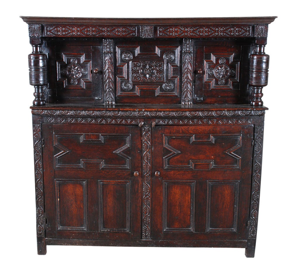 An English oak court cupboard. The geometric molded panels of the upper section carved with Tudor roses. The geometric paneled doors of the lower section surrounded by foliate carved stiles.